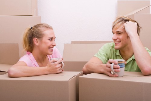 A young woman and a young man, both with light-colored hair, are sitting inside a residential space surrounded by stacked cardboard moving boxes. The woman, wearing a pink top, is smiling and holding a smartphone, while the man, dressed in a green t-shirt, is smiling back and resting his head on his hand. They are engaged in a friendly conversation amid the boxes, suggesting a recent or ongoing house move. Natural lighting fills the room, which has plain white walls, and the scene implies a casual, relaxed atmosphere during a household relocation process. The background shows more cardboard boxes stacked against the wall, indicating an active moving or packing session typical of house removals in areas like Crystal Palace or nearby London neighborhoods, with the scene representing a typical internal setting for professional house moving services.