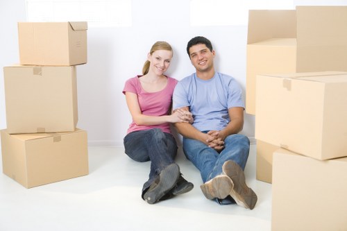 A young couple sitting on a white floor surrounded by stacked cardboard moving boxes in a bright, empty room with white walls. The woman is wearing a pink t-shirt and jeans, with blonde hair tied back, and is smiling while holding the man's hand. The man is wearing a light blue t-shirt and dark blue jeans, with short dark hair, and is also smiling. The boxes vary in size and are arranged on both sides of the couple, some stacked two or three high, indicating a house move. The room has natural lighting, providing a clear view of the couple and the packing process, which aligns with professional house removals services in the local area of Crystal Palace or nearby London suburbs. The scene captures a moment of packing or moving, emphasizing the careful handling of household belongings, suitable for a removals service website.