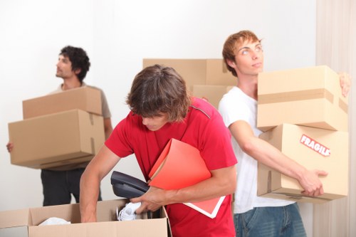 Three movers in a residential household setting, preparing for a house relocation in the Crystal Palace area. The central figure, wearing a red shirt, is leaning over a large cardboard box and holding a cordless drill and a red folder, with a black smartphone tucked under their arm. To their right, a young man dressed in a white t-shirt is carrying a brown cardboard box labeled 'FRAGILE,' with a slightly upward gaze, possibly towards the doorway or window. In the background, another team member, dressed in a grey shirt, is seen holding a stack of similar cardboard boxes, facing away from the camera. The scene takes place inside a house with a neutral-colored wall and a window with vertical blinds, indicating natural daylight filling the room. Several open cardboard boxes are visible, filled with household items, including a table lamp and clothing. All items are made of cardboard, with some boxes flipped open showing miscellaneous belongings. The movers are engaged in packing or preparing items for transportation related to a house move in the Crystal Palace or nearby postcode area. The environment appears organized and professional, emphasizing the company’s focus on careful handling during removals in south London.