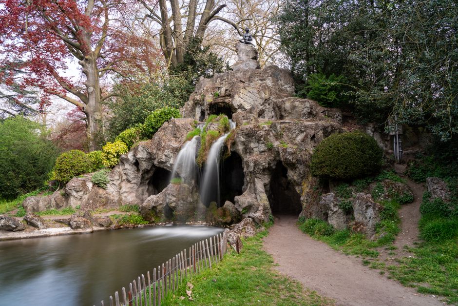 A section of a park with a decorative flower mosaic made from colorful tiles, situated on a raised platform bordered by a grey metal railing. In the background, there are three sculpted stone figures